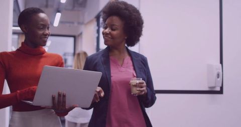 Professional women collaborating in office hallway with laptop and coffee