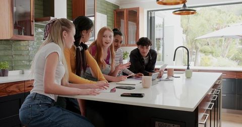 Diverse teenage group studying around sunlit kitchen island with laptops and notebooks