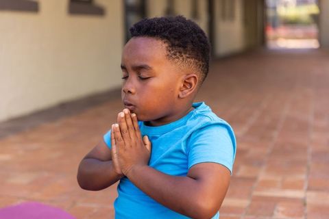 African american boy praying with closed eyes kneeling on hallway floor wearing blue shirt