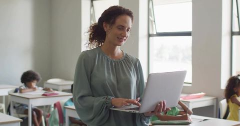 Smiling teacher teaching while using laptop in bright classroom with focused students