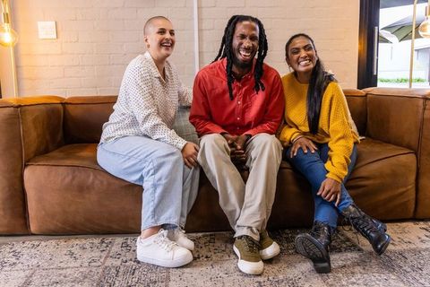 Friends laughing together on brown leather sofa in cozy lounge, casual multicultural trio