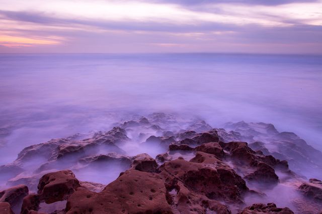Purple hues blend with the evening sky over a rocky coastal seashore, where the ocean laps gently against stones. Perfect for backgrounds, nature themes, travel posters, or serene wallpaper.