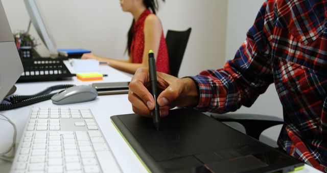 Young professionals are working in a modern office environment, using digital tools to complete tasks. One person is using a digital tablet for creative work, while another is concentrating on their computer. This visual can be used for content related to modern work environments, digital tools in professional settings, teamwork, collaboration, and the creative industry.