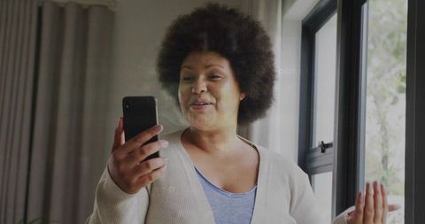 Woman video calling on smartphone while gesturing by window in cozy living room wearing cardigan