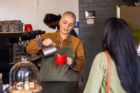 Barista pouring steamed milk into red mug while serving customer at busy neighborhood cafe