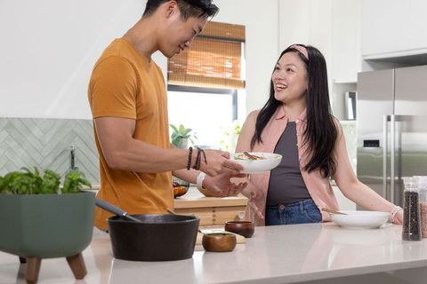 Asian couple sharing homemade salad while passing bowl at sunlit modern kitchen island