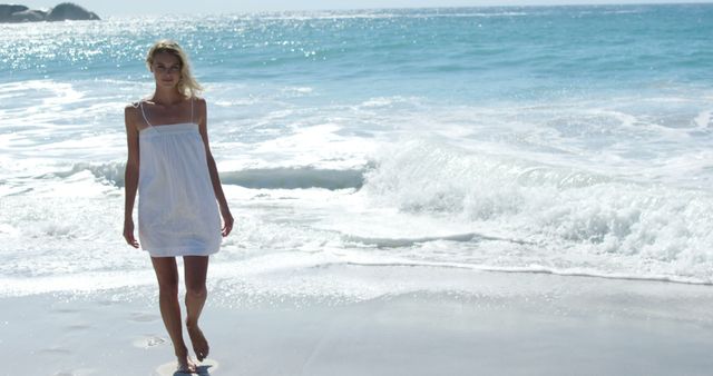 Blonde woman in white dress walking along sandy beach with gentle ocean waves in background. Perfect for themes of relaxation, summer vacations, coastal living, and leisure activities.
