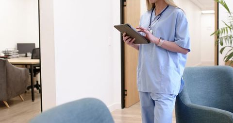 Female nurse holding tablet checking electronic records in clinic hallway, medical staff