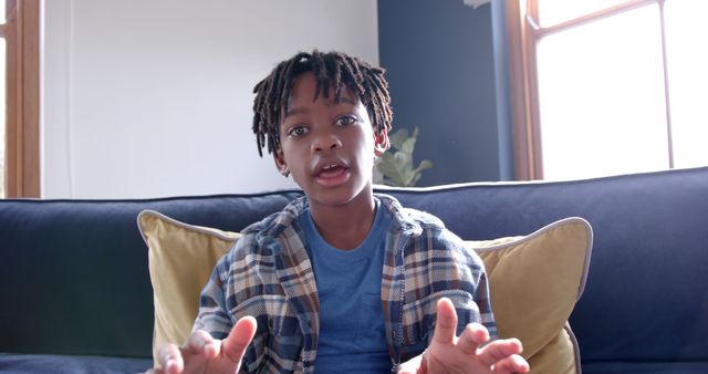 Young boy with dreadlocks sitting on a couch, talking and using hand gestures. Daytime light comes through the window behind him. He is wearing a plaid shirt over a blue t-shirt and appears to be explaining or sharing something important. Ideal for content about communication, childhood emotions, and expressive behavior.