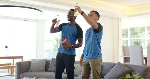 Two friends celebrating game day at home holding football and raising drinks in living room