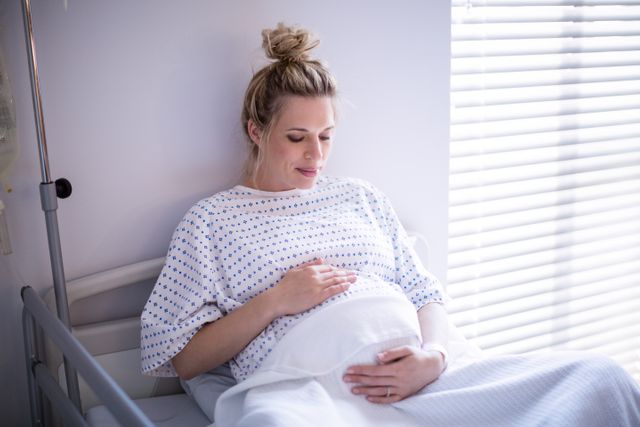 Pregnant woman relaxing on hospital bed in maternity ward, gently touching her belly. Ideal for use in healthcare, maternity, prenatal care, and hospital-related content. Can be used in brochures, websites, and educational materials about pregnancy and hospital care.