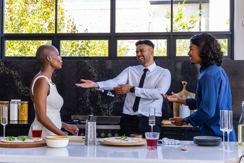 Friends laughing and serving flatbreads around modern kitchen island with champagne