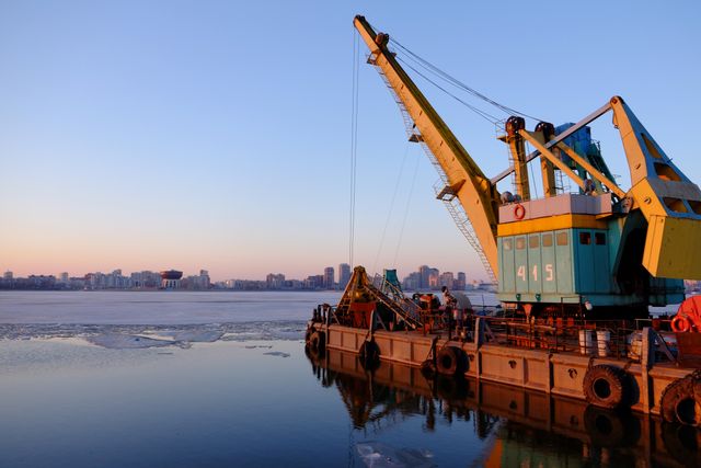 Large industrial crane on barge anchored on icy river during sunrise with city skyline in background. Ideal for use in articles about industrial work, construction, urban development, waterfront activities, engineering projects, and wintertime industrial operations.