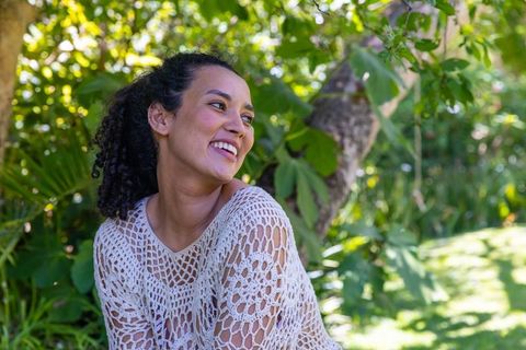 Smiling african american woman sitting by tree wearing crochet sweater in sunlit garden