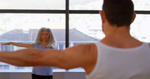 A middle-aged Caucasian woman is practicing yoga or stretching indoors, facing a young Caucasian man who appears to be a fitness instructor or personal trainer, with copy space. Their focus and alignment suggest a session of physical wellness or personal training.