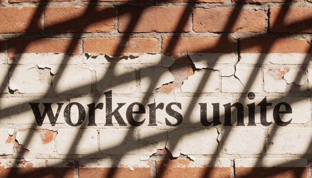Displaying painted slogan workers unite on weathered red brick wall under diagonal shadows