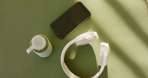 White over-ear headphones on green yoga mat with phone and water bottle, sunlit fitness
