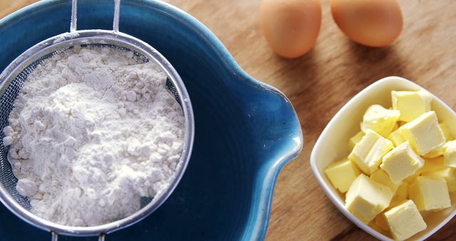 A sieve with flour is placed over a blue mixing bowl next to a dish of cubed butter and eggs on a wooden surface, with copy space. Ingredients are prepared for baking, suggesting the beginning stages of a recipe such as pastry or cake making.