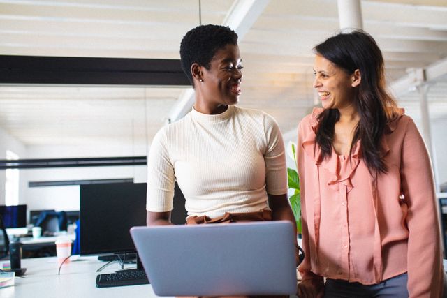 Two diverse businesswomen are collaborating in a modern office environment. They are smiling and engaging in a friendly conversation while working on a laptop. This image can be used to depict themes of teamwork, diversity, professional collaboration, and a positive work atmosphere. Ideal for business websites, corporate presentations, and articles on workplace culture.