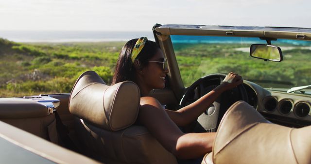 Woman enjoying a drive in a convertible car along a scenic coastal road with a beautiful landscape in the background. Suitable for travel advertisements, articles about road trips and scenic drives, lifestyle blogs, promotional materials for car companies, and summer vacation brochures.