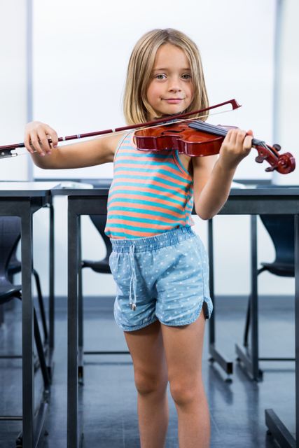 Young girl standing in classroom playing violin. She is wearing casual clothes and appears to be focused on her music. Suitable for use in educational materials, advertisements for music classes, promotional content for schools, or articles about child development and learning.