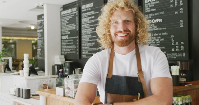 Friendly barista with curly blond hair greeted customers in modern cafe. Background shows menu boards and contemporary decor. Ideal for promoting coffee shop culture, hospitality industry, and customer service.