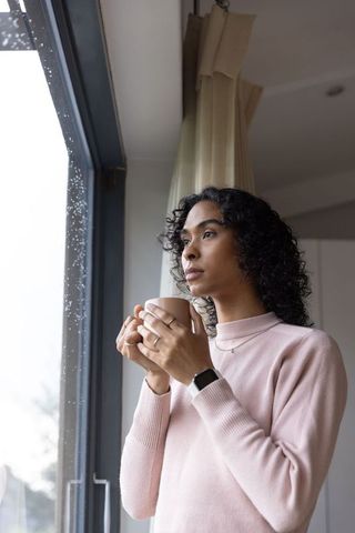 Non-binary adult standing by rainy window holding mug, wearing smartwatch, contemplative