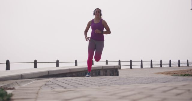 Biracial woman using headphones running on promenade by sea - Download Free Stock Photos Pikwizard.com