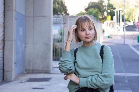 Contemplating young woman standing on urban sidewalk wearing green sweatshirt and backpack