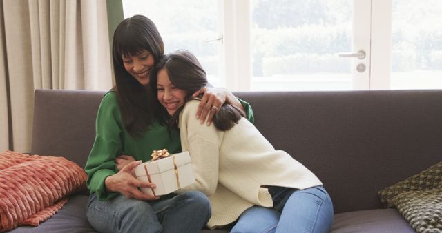 Mother and daughter hugging while holding a gift box on a sofa. Both are smiling, sharing a joyful and loving moment. Perfect for themes related to family bonding, celebrations, and special occasions. Can be used in advertisements, brochures, family-oriented publications, or websites promoting family products and services.