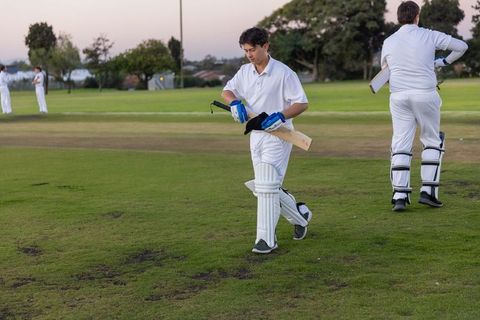 Cricket teammates walking across suburban pitch at dusk wearing white uniforms, carrying bats