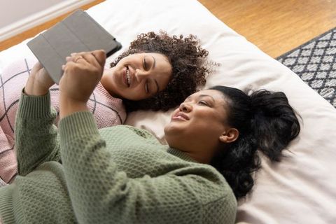 Multicultural women relaxing on bed together using tablet in cozy sweaters sharing laughs