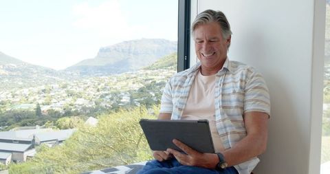 Mature man smiling and using tablet on window seat with panoramic mountain valley view