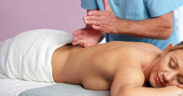 Woman lying on massage table with eyes closed receiving back massage from professional, wrapped in white towel. Ideal for promoting wellness clinics, spa services, stress relief products, and self-care practices.