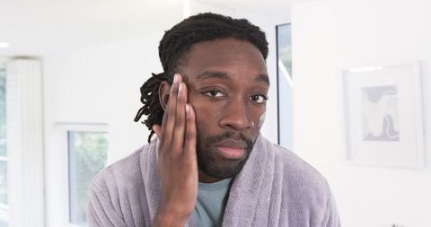 African american man applying moisturizer with nose hoop wearing purple robe and dreadlocks