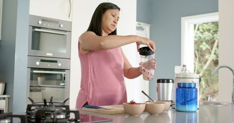 Mature woman preparing strawberry smoothie on modern kitchen island, healthy lifestyle