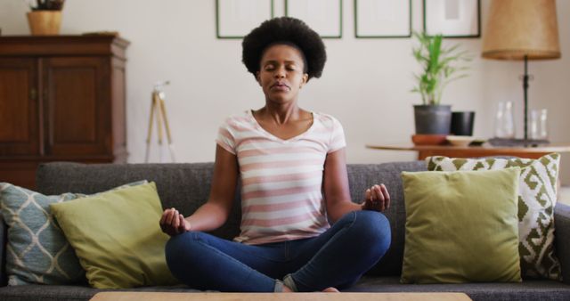 Woman sitting cross-legged on sofa, meditating and practicing yoga. She appears focused and calm, wearing casual clothes with home decor in the background. Ideal for concepts related to mental health, relaxation, mindfulness, and home wellness routines.