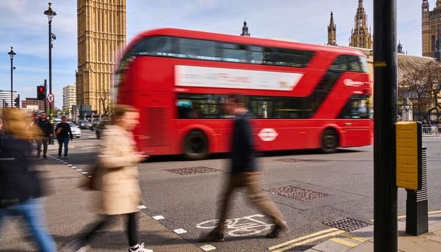 Red double-decker bus speeding past westminster and big ben with blurred commuters crossing
