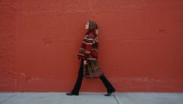 Woman walking in patterned knit hooded coat and heeled ankle boots along red urban wall