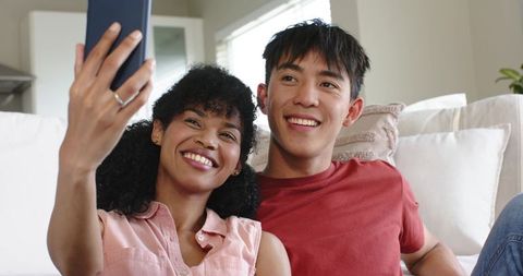Mixed-race couple taking selfie on sofa smiling and relaxing in bright modern living room