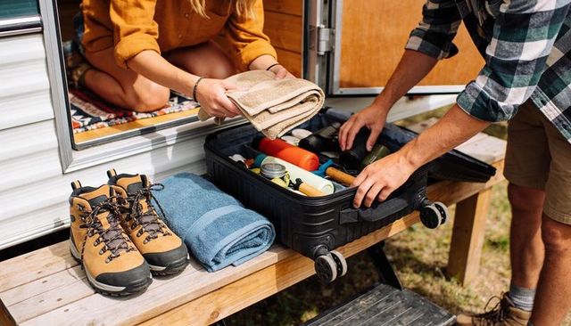Couple packing camping gear into rolling suitcase at camper door with hiking boots