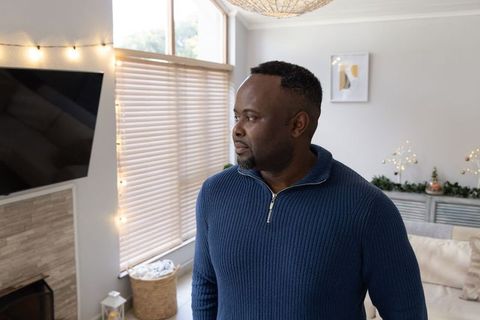 African american man standing by window in home wearing navy sweater admiring warm lights