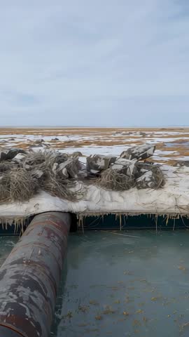 Tilting rusted pipe revealing frozen marsh embankment and rippled water vertical video