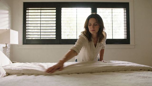 Woman straightening duvet in minimalist neutral bedroom with shuttered window morning calm