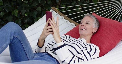 Mature woman reclining on white hammock using smartphone and smiling, backyard relaxation