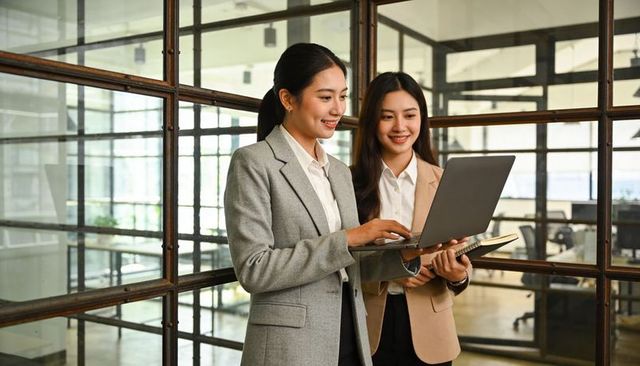 Two professional women collaborating on laptop in modern glass-partitioned office workspace