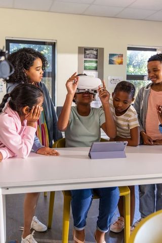 Diverse kids exploring virtual reality, child adjusting vr headset at classroom table
