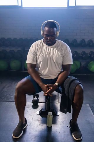 Backlit african man resting between sets wearing headphones and towel on bench with dumbbell