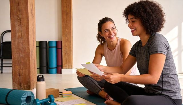Two women reviewing fitness plan in yoga studio, smiling, planning wellness routine