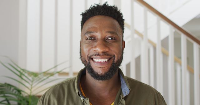 Portrait of a smiling African American man in casual clothing standing inside a home. Capture positive moments and relaxed lifestyle for use in lifestyle blogs, diversity initiatives, and personal development content.